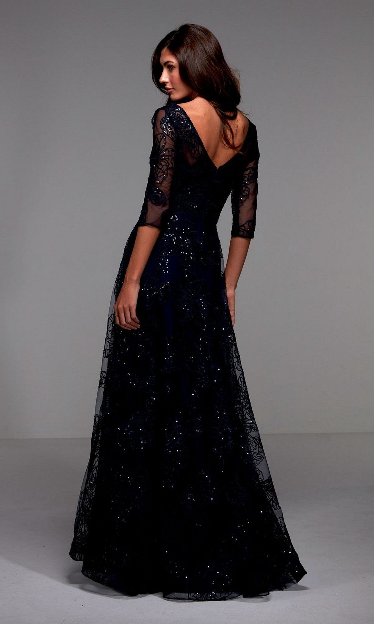 A woman wears the Alyce 27494, a long navy formal dress with sequin lace sleeves and a deep V-back, posing elegantly in a studio—perfect for the mother of the bride.